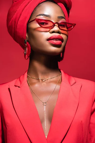 African american young woman in stylish outfit, sunglasses and turban isolated on red — Stock Photo