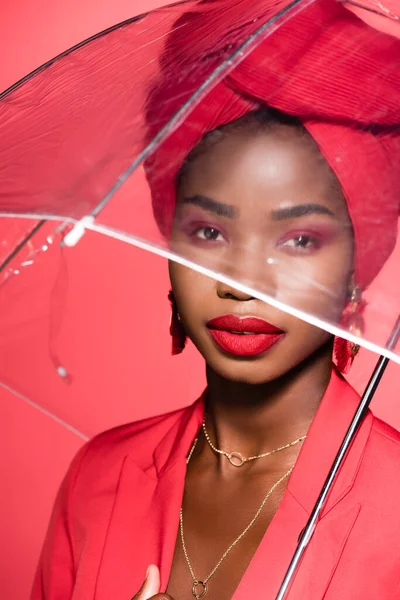 African american young woman in stylish outfit and turban holding umbrella isolated on red — Stock Photo