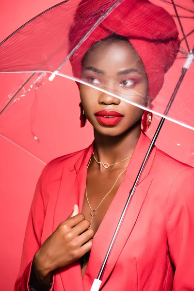 African american young woman in stylish outfit and turban holding umbrella isolated on red — Stock Photo