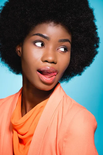 Afro americano jovem mulher em laranja elegante roupa mostrando língua isolada no azul — Fotografia de Stock