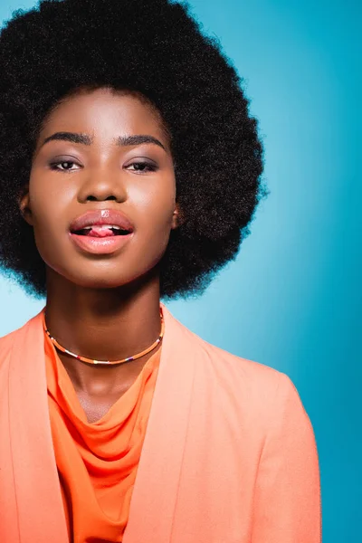 Mujer joven afroamericana en traje elegante naranja que muestra la lengua aislada en azul - foto de stock