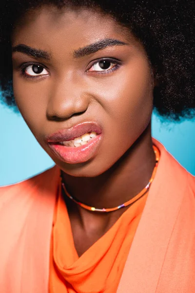 Jeune femme afro-américaine en colère dans une tenue élégante orange montrant des dents isolées sur bleu — Photo de stock