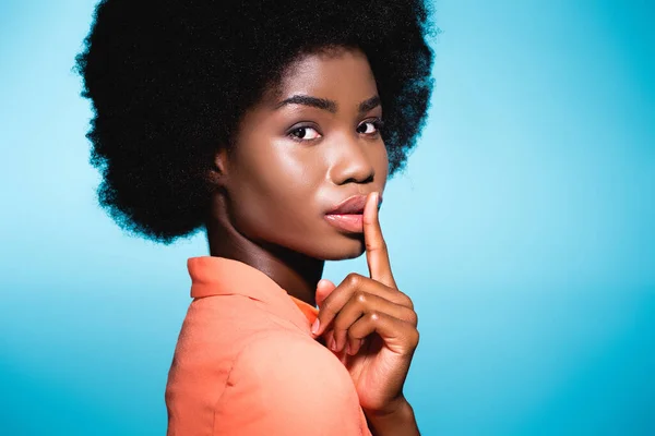 African american young woman in orange stylish outfit showing shh isolated on blue — Stock Photo