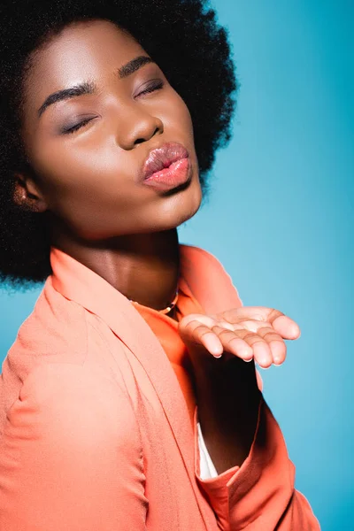 Афро-американська молода жінка в помаранчевому стильному вбранні, що дме поцілунок ізольовано на синьому — стокове фото