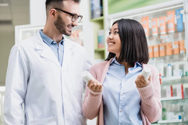 Alegre farmacéutico en gafas mirando asiático mujer sosteniendo botellas con pastillas en farmacia - foto de stock