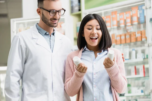 Excited asian woman holding bottles with pills near pharmacist in drugstore — Stock Photo