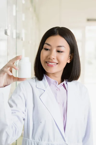 Alegre asiático farmacéutico en blanco abrigo mirando botella con medicación - foto de stock