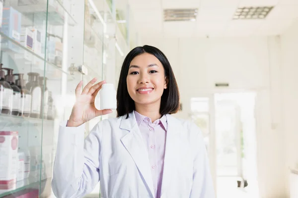 Весёлый азиатский фармацевт в белом халате, прячущий бутылку с лекарствами в аптеке — стоковое фото