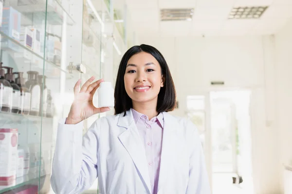 Cheerful asian pharmacist in white coat holing bottle with medication in drugstore — Stock Photo