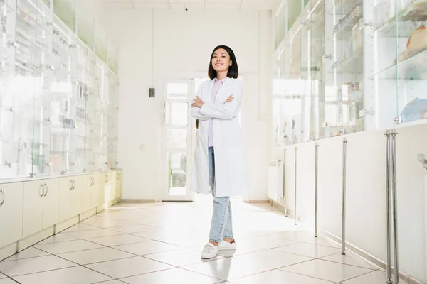 Comprimento total de sorrir asiático farmacêutico em branco casaco de pé com cruzados braços em farmácia — Fotografia de Stock
