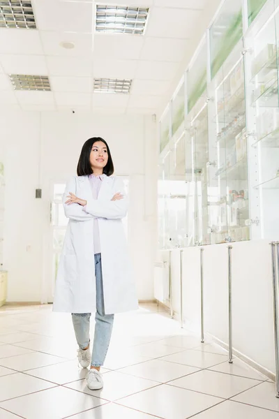 Longitud completa de sonriente asiático farmacéutico en blanco abrigo caminar con cruzado brazos en droguería - foto de stock