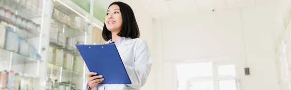 Alegre asiático farmacéutico en blanco capa celebración portapapeles en droguería, bandera - foto de stock