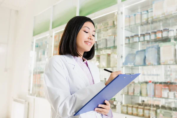 Cheerful asian pharmacist in white coat holding pen near clipboard in drugstore — Stock Photo