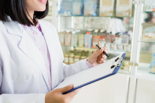 Vista recortada de farmacéutico en pluma de retención de abrigo blanco cerca del portapapeles en la farmacia - foto de stock