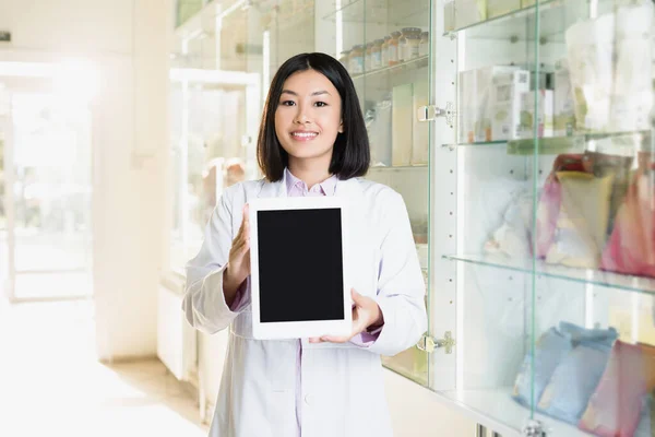 Cheerful asian pharmacist in white coat holding digital tablet with blank screen in drugstore — Stock Photo