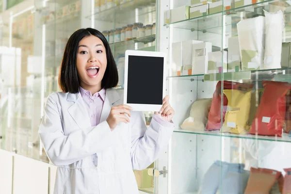 Excitado farmacéutico asiático en blanco abrigo celebración tableta digital con pantalla en blanco en farmacia - foto de stock