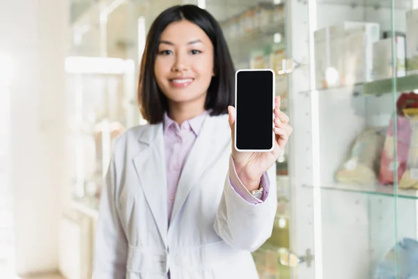 Smartphone con pantalla en blanco en la mano del alegre farmacéutico asiático en bata blanca sobre fondo borroso - foto de stock
