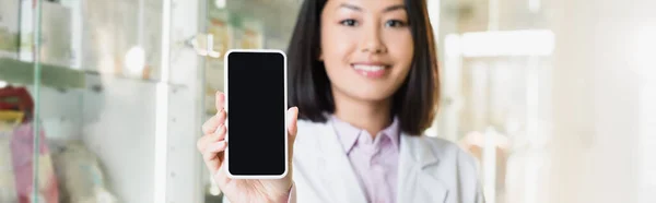 Веселый азиатский фармацевт в белом халате держит смартфон с пустым экраном в аптеке, баннер — стоковое фото