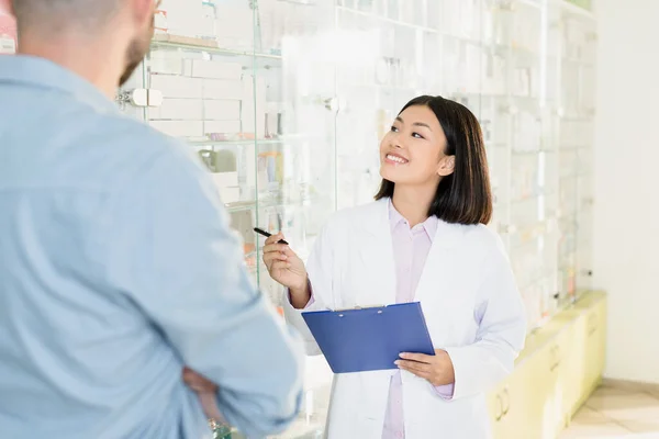 Alegre asiático farmacéutico en blanco capa celebración portapapeles cerca hombre en droguería - foto de stock