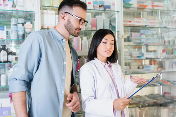 Alegre asiático farmacéutico en blanco capa apuntando con mano en portapapeles cerca hombre en droguería - foto de stock