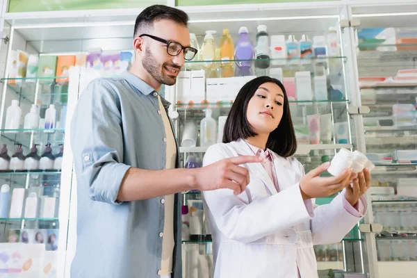 Bearded man pointing with finger at bottles with medication in hands of asian pharmacist in drugstore — Stock Photo