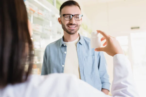 Cheerful customer looking at pharmacist holding bottle with medication on blurred foreground — Stock Photo