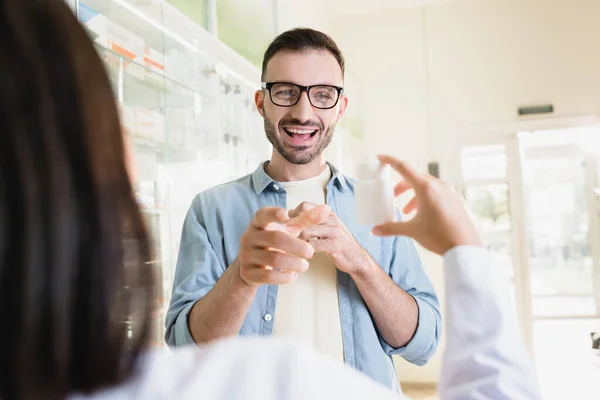 Homem animado em óculos olhando para farmacêutico segurando garrafa com medicação em primeiro plano borrado — Fotografia de Stock
