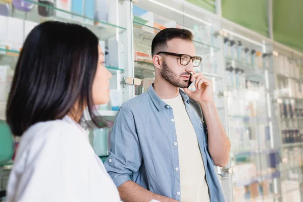 Чоловік в окулярах говорить на смартфоні біля азіатського фармацевта на розмитому фоні — стокове фото