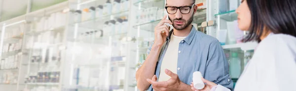 Client en lunettes parlant sur smartphone près d'un pharmacien asiatique avec bouteille de médicament au premier plan flou, bannière — Photo de stock