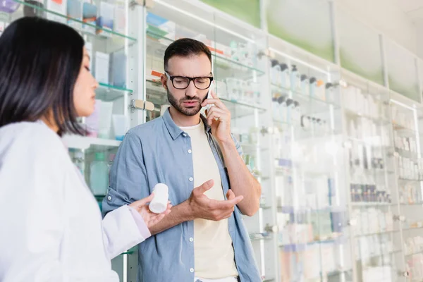 Kunde in Brille unterhält sich mit Smartphone in der Nähe eines asiatischen Apothekers mit Medikamentenflasche im verschwommenen Vordergrund — Stockfoto