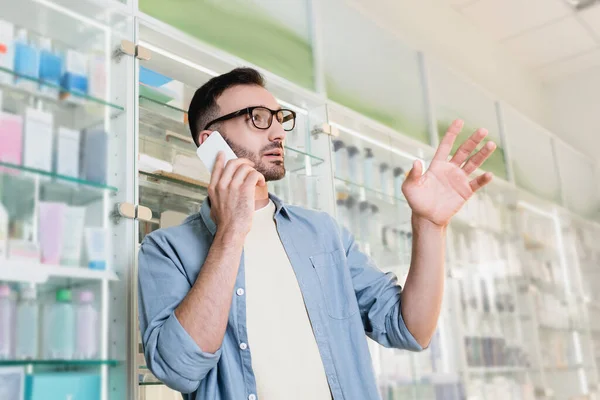 Mann mit Brille spricht auf Smartphone und gestikuliert in Drogerie — Stockfoto
