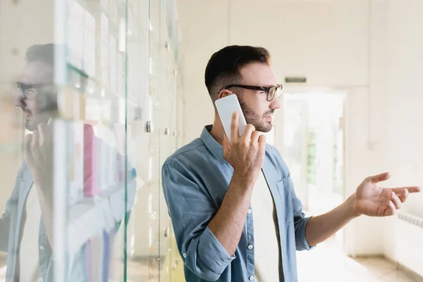 Side view of man in eyeglasses talking on smartphone while pointing with finger in drugstore — Stock Photo