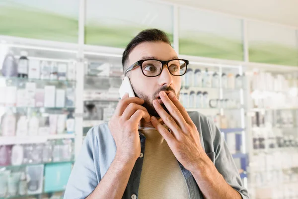 Homem chocado em óculos falando no smartphone enquanto cobre a boca na farmácia — Fotografia de Stock