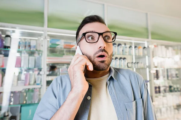 Geschockter Mann mit Brille telefoniert in Drogerie mit Smartphone — Stockfoto