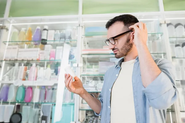 Thoughtful man in eyeglasses looking at bottle with pills in drugstore — Stock Photo