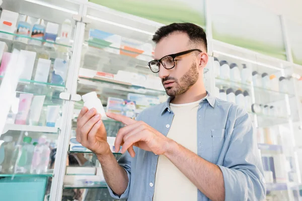 Uomo in occhiali che punta con il dito alla bottiglia con le pillole durante la lettura di prescrizione in farmacia — Foto stock