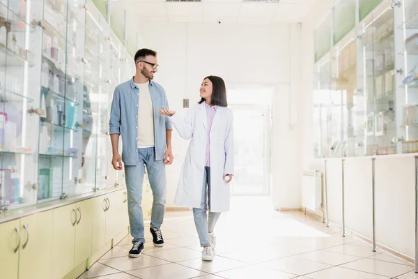 Полная длина веселый азиатский фармацевт в белом халате ходьба с клиентом в аптеке — стоковое фото