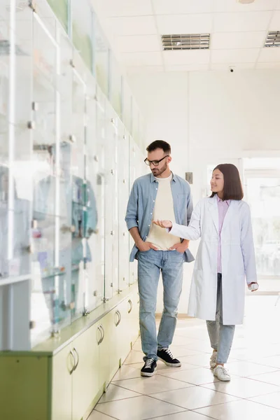 Full length of cheerful asian pharmacist in white coat pointing with hand while showing medication to man — Stock Photo