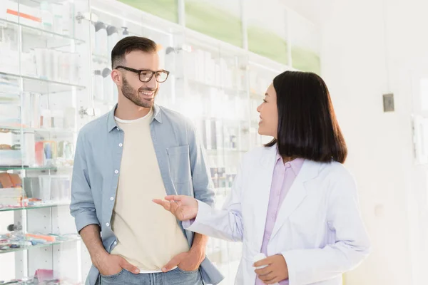 Cheerful asian pharmacist in white coat pointing with finger while talking with customer — Stock Photo