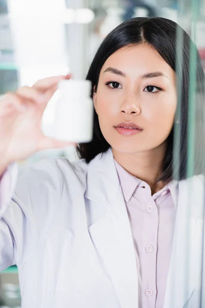 Задумчивый азиатский фармацевт в белом халате смотрит на бутылку на размытом переднем плане — стоковое фото