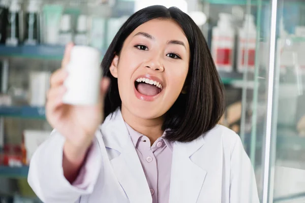Возбужденный азиатский фармацевт в белом халате держит бутылку с лекарствами на размытом переднем плане — стоковое фото