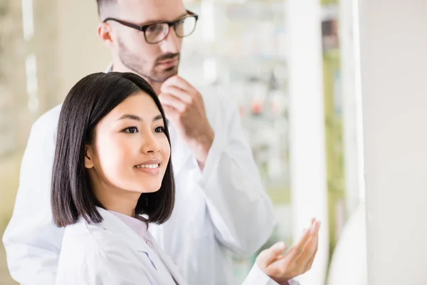 Smiling asian pharmacist in white coat pointing with hand near colleague in eyeglasses on blurred background — Stock Photo