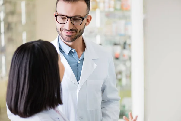 Smiling pharmacist in white coat and eyeglasses looking at asian colleague on blurred foreground — Stock Photo