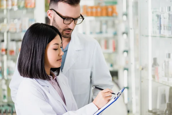 Interracial pharmacists in white coats looking at clipboard in drugstore — Stock Photo