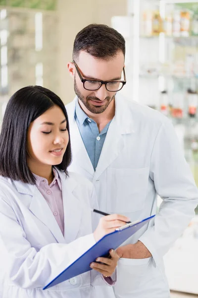 Smiling interracial pharmacists in white coats looking at clipboard in drugstore — Stock Photo