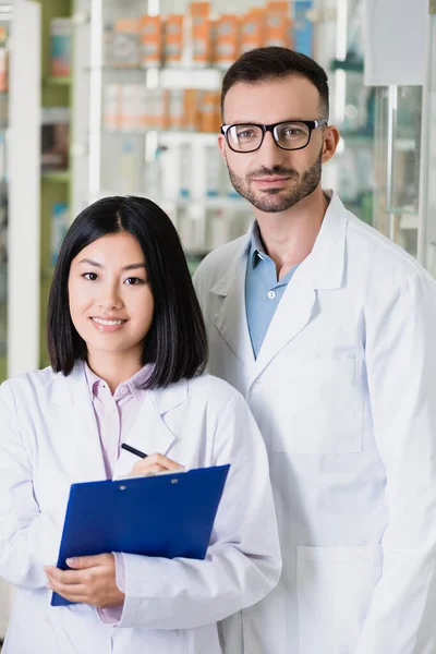 Pharmaciens interracial souriants en manteaux blancs regardant la caméra en pharmacie — Photo de stock