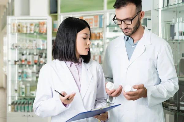 Farmacêuticos inter-raciais em casacos brancos olhando para garrafas com medicação na farmácia — Fotografia de Stock