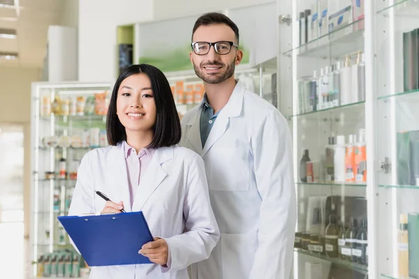 Happy interracial pharmacists in white coats looking at camera in drugstore — Stock Photo