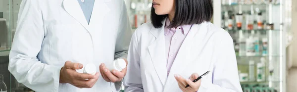 Vue recadrée du pharmacien en manteau blanc tenant des bouteilles avec des médicaments près de son collègue en pharmacie, bannière — Photo de stock