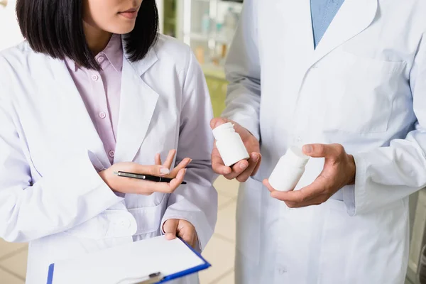 Partial view of pharmacist in white coat holding bottles with medication near colleague in drugstore — Stock Photo